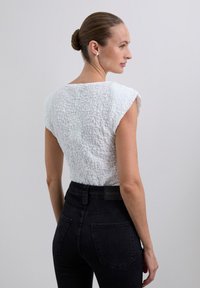 Weißes, strukturiertes Oberteil mit kurzen Ärmeln, kombiniert mit schwarzen, hoch taillierten Jeans. Das Oberteil weist dezente, zerknitterte Details auf.