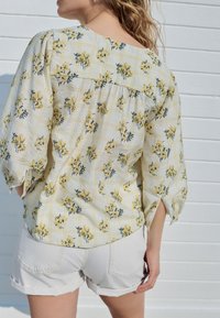 Blusa floreale giallo chiaro con motivo a griglia, maniche a tre quarti e dettaglio plissettato sul retro, abbinata a pantaloncini bianchi.