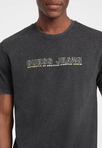 Pilkas medvilninis marškinėliai su užrašu "GUESS JEANS" ryškiomis juodomis ir geltonomis raidėmis. Klasikinis apvalus kaklas ir trumpomis rankovėmis.