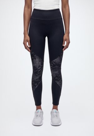 Un mannequin se tient debout face à l’avant, vêtu d’un legging de sport noir avec des empiècements à motifs aux genoux, d’un haut blanc et de baskets blanches.