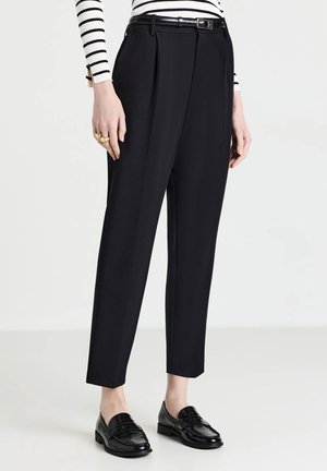 Frau trägt schwarze maßgeschneiderte, knöchellange Hose mit schwarzem Gürtel, schwarzen Slippern und einem weißen Pullover mit schwarzen horizontalen Streifen.
