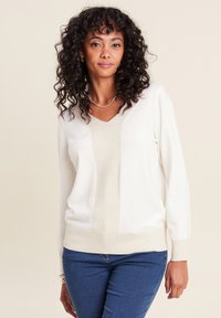 Maglione crema chiaro con scollo a V e centro texturizzato, dotato di maniche lunghe e una vita aderente. Abbinato a jeans blu.