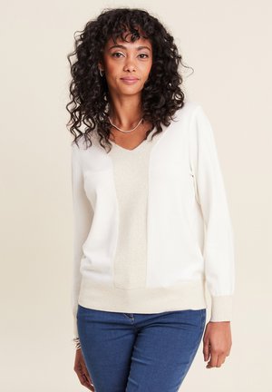 Hellbeiger Pullover mit V-Ausschnitt und strukturiertem Zentrum, mit langen Ärmeln und einem taillierten Bündchen. Kombiniert mit blauen Jeans.