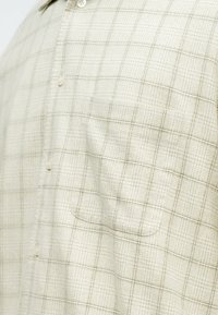 Chemise à carreaux beige clair avec un motif quadrillé subtil, une poche avant et de petits boutons au centre.