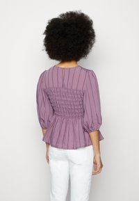 Femme aux cheveux bouclés portant un chemisier froncé violet à manches bouffantes trois-quarts et un pantalon blanc, debout face à un fond blanc.