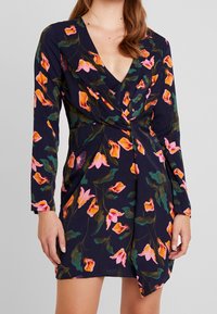 Sukienka w kwiaty z dekoltem w serek, długimi rękawami i granatowym tłem z żywymi różowymi i zielonymi wzorami kwiatowymi.