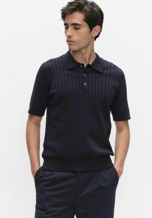 Mężczyzna ubrany w granatową, prążkowaną koszulkę polo z krótkim rękawem oraz dopasowane spodnie, z rękami w kieszeniach, patrzący w bok na jednolitym tle.