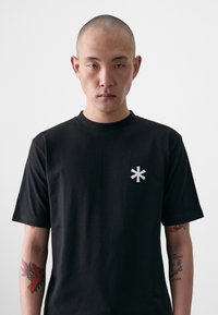 Black cotton t-shirt with a round neck, short sleeves, and a white asterisk logo on the left chest. Smooth texture, fitted design.