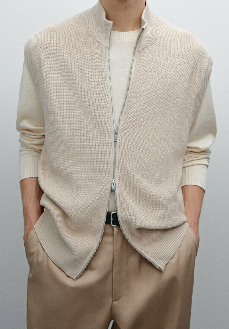 Beiger ärmelloser Reißverschluss-Pullover mit geripptem Muster, hohem Kragen und Seitentaschen, kombiniert mit einem cremefarbenen Langarmshirt.