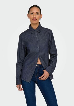 Camisa de mezclilla azul oscuro con collar puntiagudo, mangas largas y cierre de botones. Presenta un corte recto y un frente ligeramente abierto.