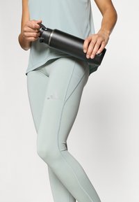 Čierna fľaša na vodu z nehrdzavejúcej ocele s výpustom, držená v ruke, vedľa svetlozelených legín so zvýrazneným prešitím a logom.