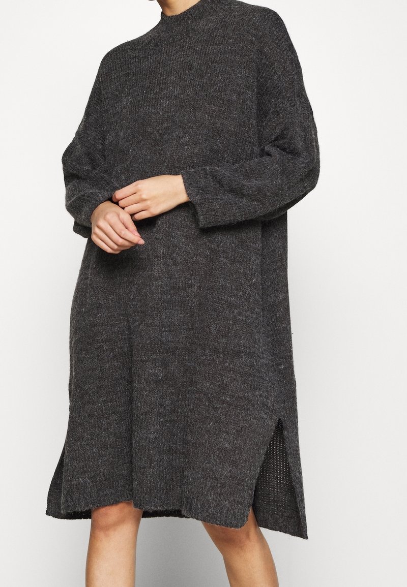 Personne portant une robe pull en tricot gris foncé, ample, à manches longues et avec des fentes latérales, se tenant devant un fond clair uni.