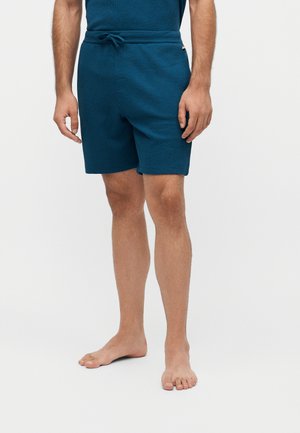 Homme portant un short bleu sarcelle avec cordon et une chemise assortie, debout pieds nus devant un fond blanc uni.