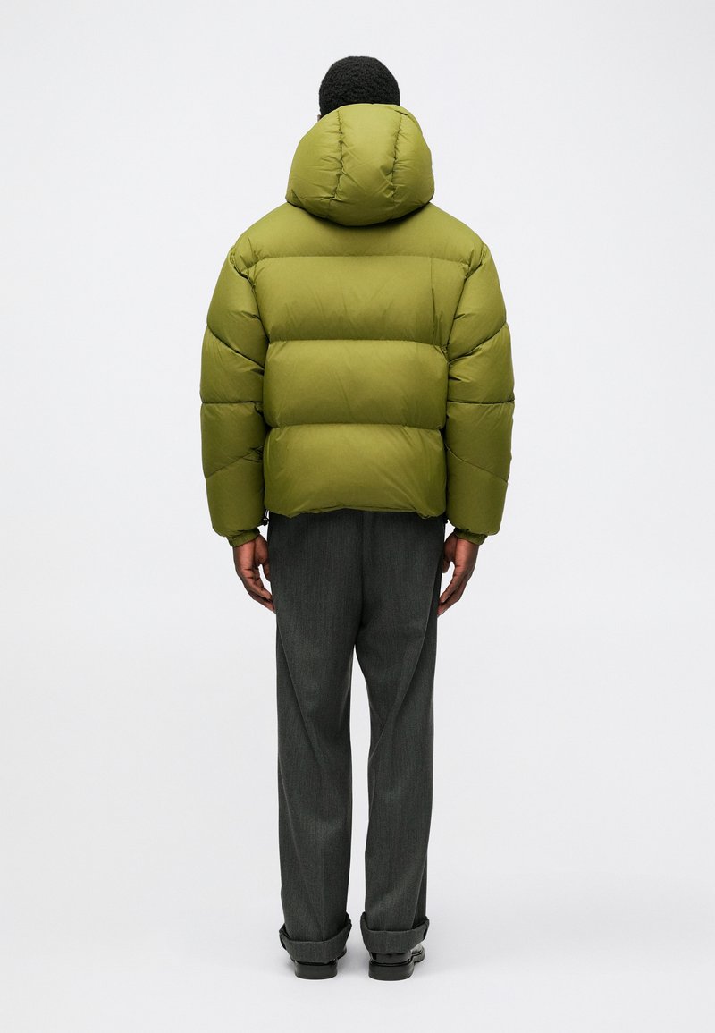 Olive green puffer jacket with a detachable hood, featuring quilted sections, worn over gray plaid trousers and black shoes.