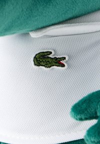 Πράσινο κεντημένο λογότυπο κροκοδείλου σε λευκό υφασμάτινο φόντο με υφή, με πράσινο υλικό από βελούδο ορατό στις άκρες.