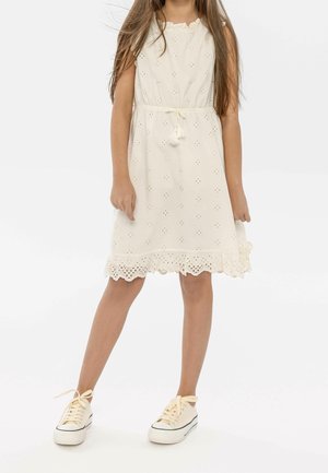 White embroidered dress with a scalloped hem, drawstring waist, and tassel details, paired with light-colored sneakers.