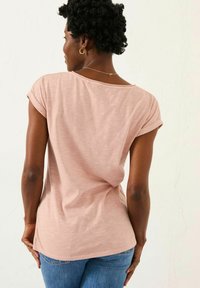 Light pink short-sleeve top with a scoop neckline, made of soft fabric. Features rolled sleeves and a loose fit, paired with blue jeans.