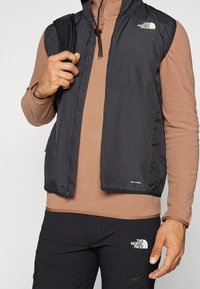 Gilet antivento nero con cappuccio, zip frontale e dettagli con logo. Indossato sopra una camicia a maniche lunghe marrone chiaro e pantaloni scuri.