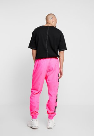 Man facing away wearing a black oversized T-shirt, bright pink joggers with side text, white sneakers, and visible neck and head tattoos.
