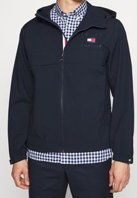 Giacca soft-shell blu navy con zip frontale, cappuccio regolabile e due tasche laterali. Presenta il logo sul petto e polsini elasticizzati.