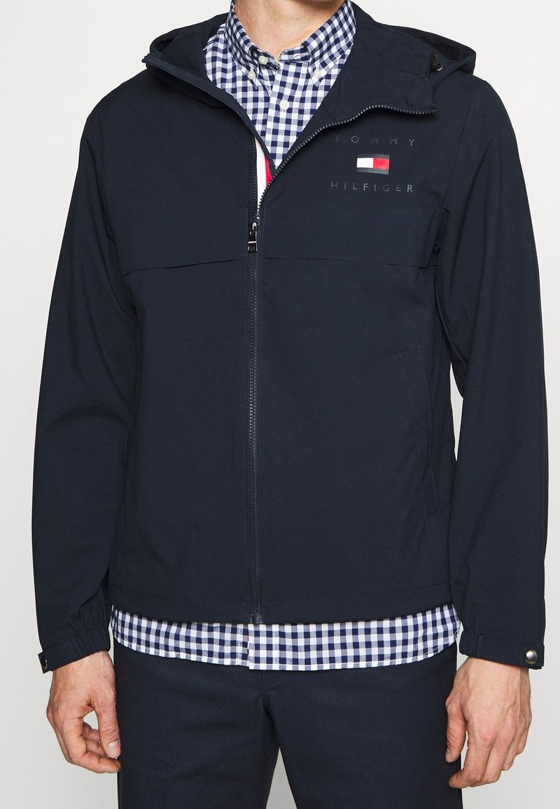 Giacca soft-shell blu navy con zip frontale, cappuccio regolabile e due tasche laterali. Presenta il logo sul petto e polsini elasticizzati.