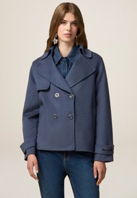 Cappotto in lana doppiopetto navy con ampi risvolti, tasche frontali e polsini con bottoni, indossato sopra una camicia di denim.