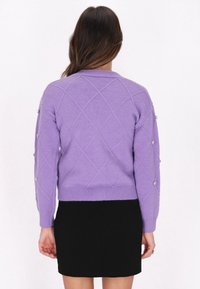 Hellpurpurner Strickpullover mit einem Diamantmuster, dekorativen Knöpfen an den Ärmeln und einer taillierten Passform, kombiniert mit einem schwarzen Rock.