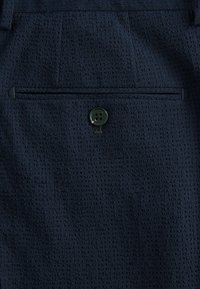 Tissu texturé bleu marine avec un petit motif en relief. Présente une poche arrière avec un bouton vert.