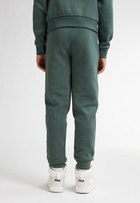 Zelené tepláky vyrobené z měkké, silné látky, mají elastické manžety a pas, doplněné bílými vysokými teniskami.