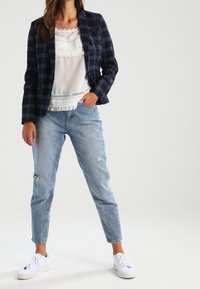 Femme portant un blazer à carreaux bleu marine, un chemisier blanc à volants, un jean clair déchiré et des baskets blanches, debout avec une main dans la poche.