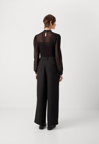Zwarte doorschijnende blouse met opstaande mouwen, kantaccenten bij de halslijn en een keyhole achterdetail, gecombineerd met wijde zwarte broeken.