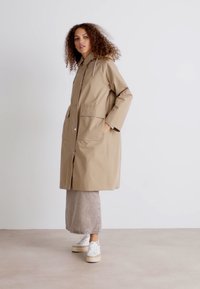 Beige waterdichte jas met een capuchon, knopen aan de voorkant en twee grote zakken. Gecombineerd met een lange spijkerrok en witte sneakers.