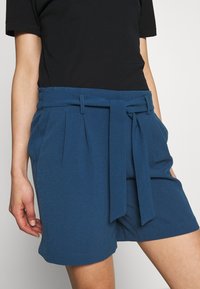 Modré texturované šortky s širokým pasem a látkovým páskem, se sklady v přední části a uvolněným střihem. V kombinaci s černým topem.