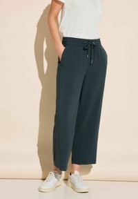 Osoba w ciemnoniebieskich spodniach z troczkiem i białych trampkach stoi oparta o beżową ścianę, z rękami w kieszeniach.