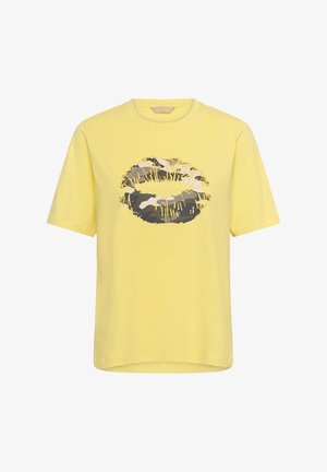 T-shirt jaune à manches courtes avec un grand motif de lèvres à motif camouflage en noir, blanc et gris, avec des accents de gouttes de peinture jaune à l'avant.