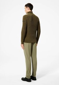 Maglione a coste verde oliva con un colletto alto, dotato di una toppa nera con logo sulla manica, abbinato a pantaloni verde chiaro e scarpe nere.