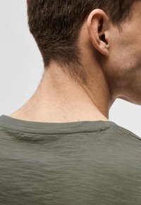 T-shirt en coton vert olive avec une coupe décontractée. Présente une large encolure et une texture subtile. Échancré près du cou et de l'épaule.