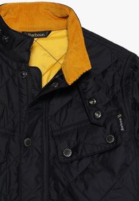 Schwarze gesteppte Jacke mit einem Kordkragen in senfgelb, Druckknöpfen und zwei Fronttaschen. Innenfutter aus glattem Stoff.