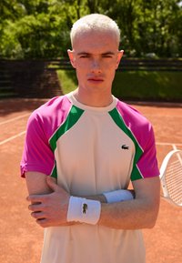 Tennis shirt with pink and green accents, short sleeves, and white body. White wristbands feature a small logo. Background shows a clay court.