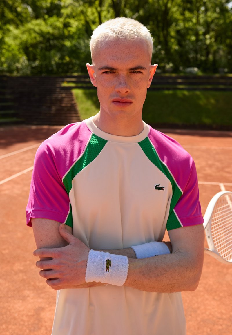 Tennistrøye med rosa og grønne detaljer, korte ermer og hvit kropp. Hvite håndleddsband har en liten logo. Bakgrunnen viser en leirbane.