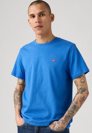 Vyras su tatuiruotėmis ant abiejų rankų, vilkintis mėlyną Levi's marškinėlį ir džinsus, stovi su rankomis kišenėse prieš neutralią fono.