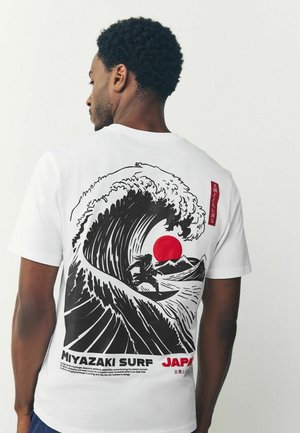 Man wearing white t-shirt with black and red graphic of surfer riding large wave with red sun and mountains, text "Miyazaki Surf Japan."