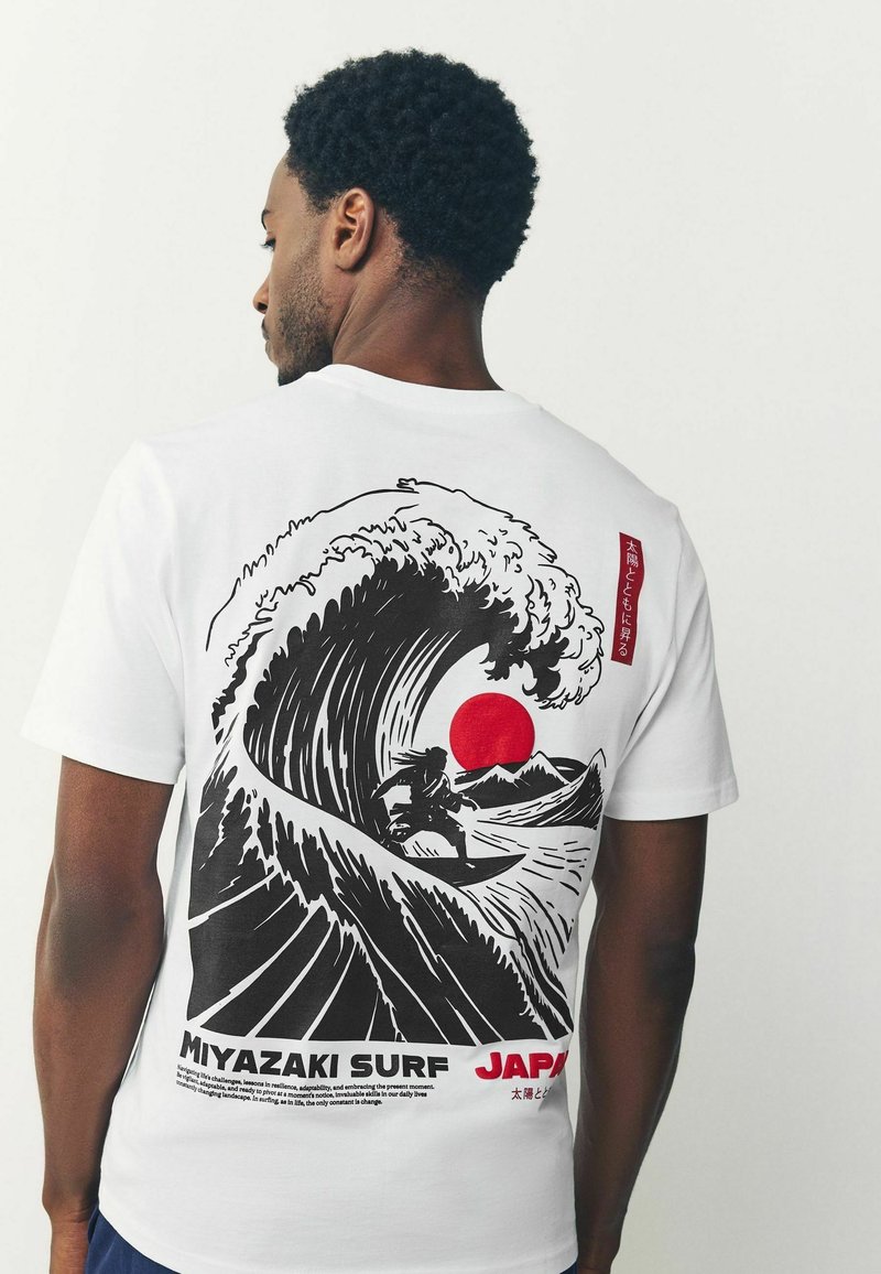 Muž v bielom tričku s čiernobielou a červenou grafikou surfistu jazdiaceho na veľkej vlne s červeným slnkom a horami, nápis "Miyazaki Surf Japan."
