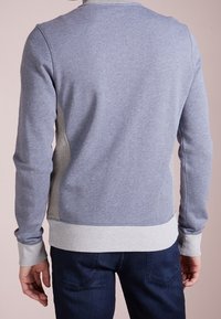 Uomo che indossa una felpa a maniche lunghe di colore azzurro chiaro e grigio e jeans blu scuro, in piedi con la schiena rivolta verso la fotocamera.