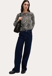 Leopardmönstrad långärmad topp i kombination med mörkblå vida jeans. Svart handväska, svarta skor, modell som står mot en ljus bakgrund.