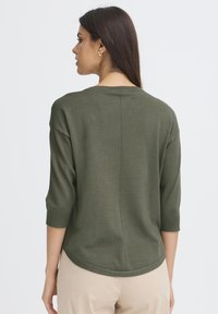 Femme aux longs cheveux noirs portant des boucles d'oreilles en or et un haut olive à manches trois-quarts, de dos contre un fond uni.