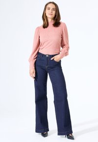 Hellrosa, strukturiertes Strickoberteil mit hohem Kragen; kombiniert mit dunkelblauen, weiten Jeans mit Kontrastnähten. Schwarze spitze Schuhe.