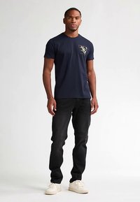 Camiseta de algodón azul marino con mangas cortas, con un gráfico floral dorado en el pecho izquierdo, combinada con vaqueros negros y zapatillas blancas.