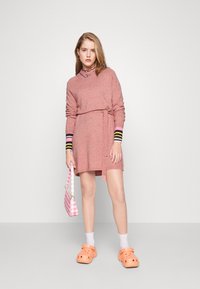 Πλεκτό ροζ φόρεμα με μακριά μανίκια, λεπτομέρεια ζώνης και ρίγες στις μανσέτες. Συνδυάζεται με πορτοκαλί καλτσάκια και τσάντα σε ροζ καρό.