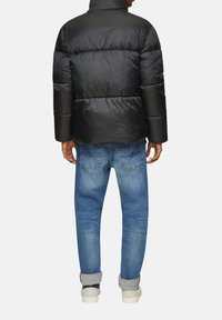 Zwarte pufferjack met horizontaal gewatteerde secties, hoge kraag, gecombineerd met lichtblauwe jeans en witte sneakers, gezien vanaf de achterkant.